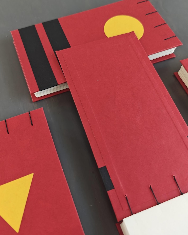 K&ouml;nnte beinhalten: Mehrere rechteckige Notizb&uuml;cher mit roten Einb&auml;nden, die schwarze Streifen und gelbe geometrische Formen wie einen Kreis und ein Dreieck aufweisen. Die Notizb&uuml;cher sind mit sichtbaren schwarzen F&auml;den an den R&auml;ndern gebunden und enthalten wei&szlig;e Papierseiten.