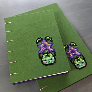 K&ouml;nnte beinhalten: Zwei gr&uuml;ne, mit Stoff bezogene Notizb&uuml;cher mit rosa N&auml;hten. Jedes Notizbuch hat ein Kreuzstichdesign einer gr&uuml;nen Kreatur mit einem lila und gelben Kopf. Die Notizb&uuml;cher sind auf einer grauen Oberfl&auml;che gestapelt.
