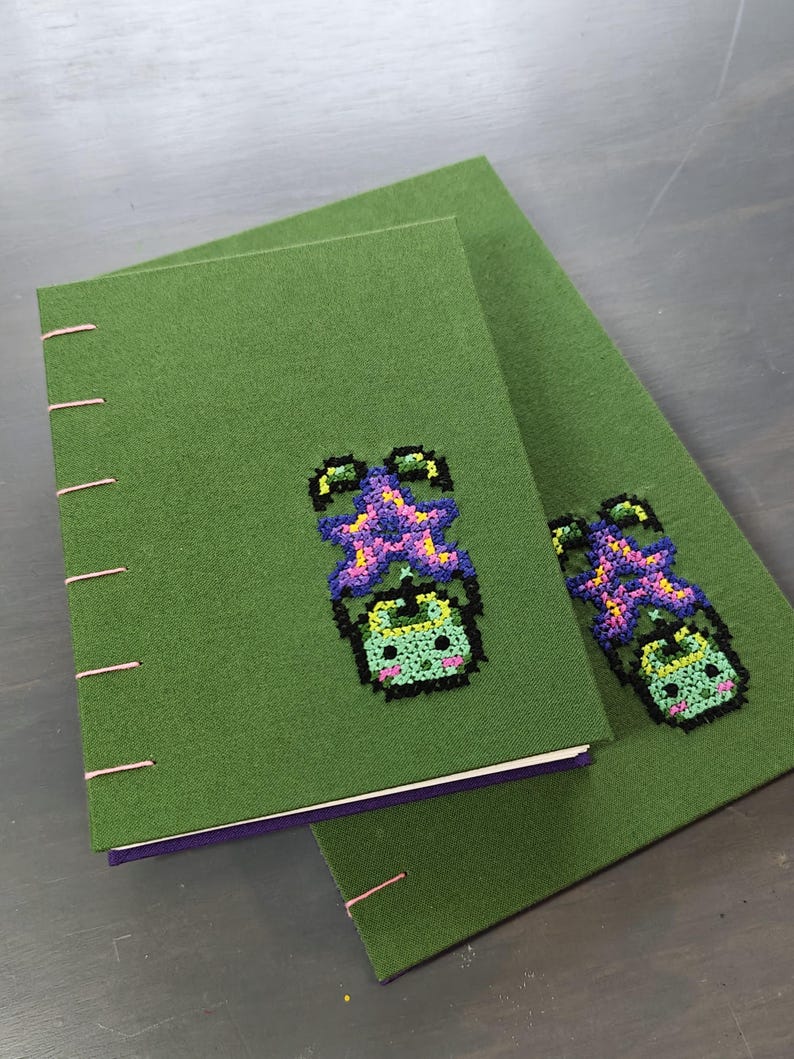 K&ouml;nnte beinhalten: Zwei gr&uuml;ne, mit Stoff bezogene Notizb&uuml;cher mit rosa N&auml;hten. Jedes Notizbuch hat ein Kreuzstichdesign einer gr&uuml;nen Kreatur mit einem lila und gelben Kopf. Die Notizb&uuml;cher sind auf einer grauen Oberfl&auml;che gestapelt.