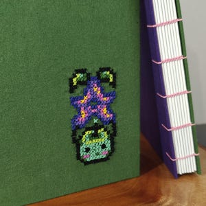 K&ouml;nnte beinhalten: Ein handgefertigtes gr&uuml;nes Notizbuch mit einem Kreuzstich-Design eines Cartoon-Charakters. Das Notizbuch hat einen lila Buchr&uuml;cken und rosa N&auml;hte. Die Seiten sind wei&szlig;. Das Notizbuch liegt auf einer Holzoberfl&auml;che.
