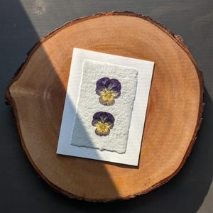 May include: A rectangular piece of off-white handmade paper with a deckled edge, featuring two pressed pansy flowers arranged vertically. The flowers have deep purple petals with yellow centers. The paper is centered on a round wooden slice.