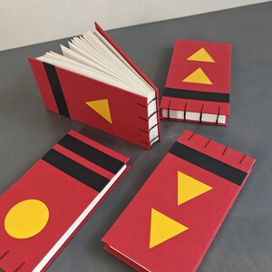 K&ouml;nnte beinhalten: Vier kleine rechteckige Notizb&uuml;cher mit roten Einb&auml;nden, schwarzen horizontalen Streifen und gelben geometrischen Formen wie Dreiecken und einem Kreis. Die B&uuml;cher haben offene wei&szlig;e Seiten und eine sichtbare schwarze Fadenheftung am Buchr&uuml;cken.