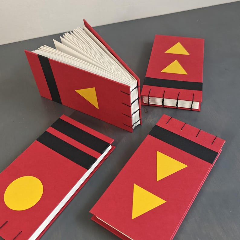 K&ouml;nnte beinhalten: Vier kleine rechteckige Notizb&uuml;cher mit roten Einb&auml;nden, schwarzen horizontalen Streifen und gelben geometrischen Formen wie Dreiecken und einem Kreis. Die B&uuml;cher haben offene wei&szlig;e Seiten und eine sichtbare schwarze Fadenheftung am Buchr&uuml;cken.