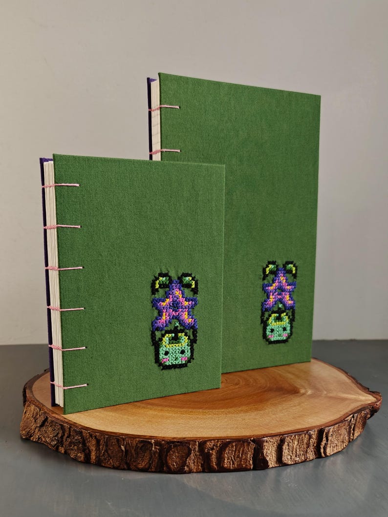 K&ouml;nnte beinhalten: Zwei gr&uuml;ne, handgefertigte Notizb&uuml;cher mit handgen&auml;hter Bindung. Jedes Notizbuch hat ein Kreuzstich-Design einer lila und gr&uuml;nen Kreatur. Die Notizb&uuml;cher werden auf einer Holzscheibe pr&auml;sentiert.