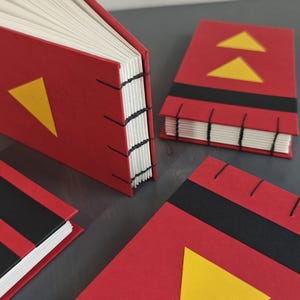 K&ouml;nnte beinhalten: Drei kleine handgefertigte B&uuml;cher mit roten Einb&auml;nden, schwarzen horizontalen Streifen und gelben Dreiecksakzenten. Die B&uuml;cher verf&uuml;gen &uuml;ber eine sichtbare schwarze Fadenheftung am Buchr&uuml;cken, die die wei&szlig;en Papierseiten mit dem Einband verbindet.