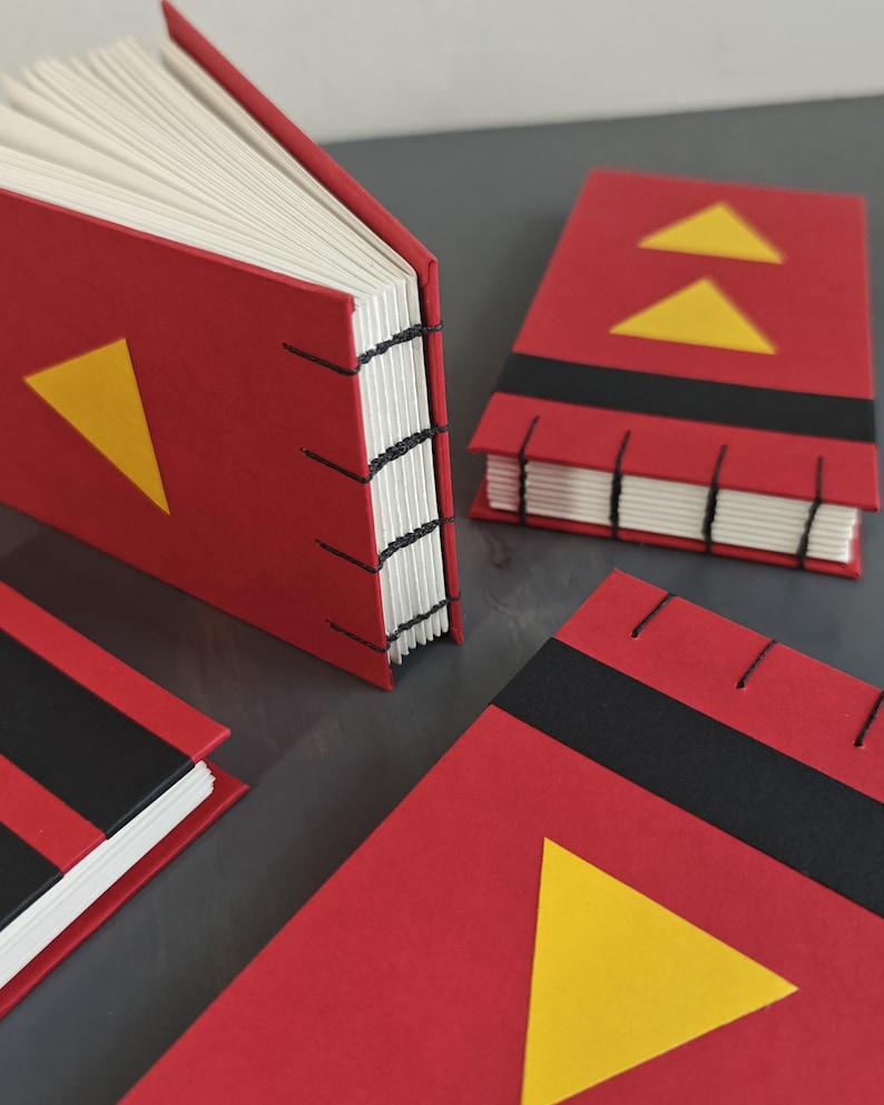 K&ouml;nnte beinhalten: Drei kleine handgefertigte B&uuml;cher mit roten Einb&auml;nden, schwarzen horizontalen Streifen und gelben Dreiecksakzenten. Die B&uuml;cher verf&uuml;gen &uuml;ber eine sichtbare schwarze Fadenheftung am Buchr&uuml;cken, die die wei&szlig;en Papierseiten mit dem Einband verbindet.