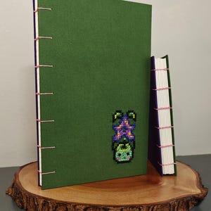 K&ouml;nnte beinhalten: Zwei handgefertigte Journale mit gr&uuml;nen Einb&auml;nden und rosa N&auml;hten. Das gr&ouml;&szlig;ere Journal zeigt ein Kreuzstichdesign einer Pflanze. Die Journale werden auf einer Holzscheibe pr&auml;sentiert.