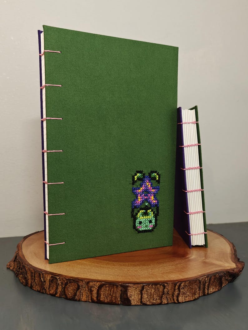 K&ouml;nnte beinhalten: Zwei handgefertigte Journale mit gr&uuml;nen Einb&auml;nden und rosa N&auml;hten. Das gr&ouml;&szlig;ere Journal zeigt ein Kreuzstichdesign einer Pflanze. Die Journale werden auf einer Holzscheibe pr&auml;sentiert.