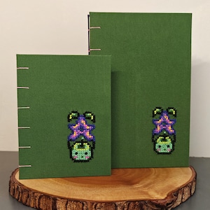 K&ouml;nnte beinhalten: Zwei gr&uuml;ne Notizb&uuml;cher mit kupferfarbener Bindung und einem kreuzgestickten Fledermausdesign. Die Fledermaus hat einen lila seesternartigen Kopf und einen gr&uuml;nen K&ouml;rper. Die Notizb&uuml;cher werden auf einer Holzscheibe pr&auml;sentiert.