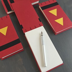 K&ouml;nnte beinhalten: Drei rote rechteckige Notizb&uuml;cher mit schwarzen und gelben geometrischen Akzenten, darunter Dreiecke und Kreise. Ein Notizbuch ist ge&ouml;ffnet und zeigt wei&szlig;e Seiten, auf denen ein silberner F&uuml;llfederhalter liegt. Die Einb&auml;nde haben schwarze N&auml;hte.