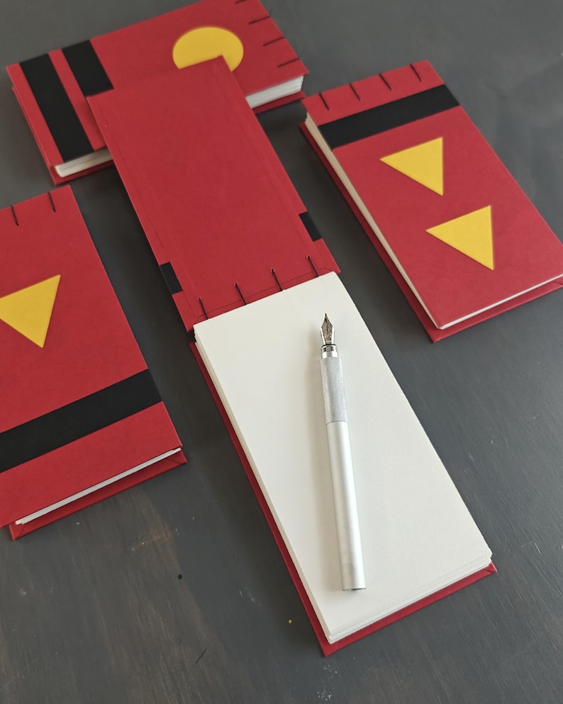 K&ouml;nnte beinhalten: Drei rote rechteckige Notizb&uuml;cher mit schwarzen und gelben geometrischen Akzenten, darunter Dreiecke und Kreise. Ein Notizbuch ist ge&ouml;ffnet und zeigt wei&szlig;e Seiten, auf denen ein silberner F&uuml;llfederhalter liegt. Die Einb&auml;nde haben schwarze N&auml;hte.