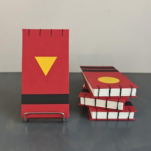 K&ouml;nnte beinhalten: Eine Sammlung kleiner, rechteckiger Notizb&uuml;cher mit roten Einb&auml;nden, schwarzen Querstreifen und sichtbaren, schwarz gen&auml;hten Buchr&uuml;cken. Ein Notizbuch steht aufrecht mit einem gelben Dreieck, w&auml;hrend andere mit einem gelben Kreis gestapelt sind.