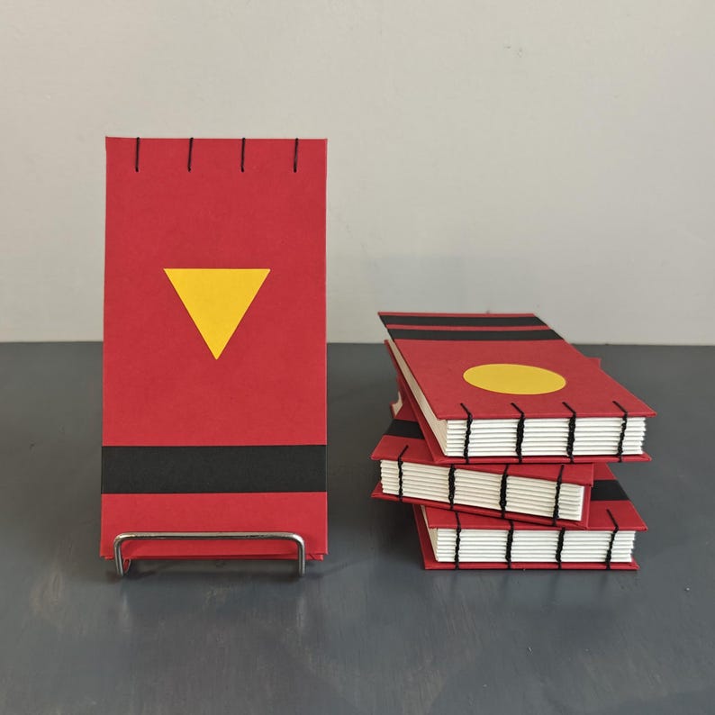 K&ouml;nnte beinhalten: Eine Sammlung kleiner, rechteckiger Notizb&uuml;cher mit roten Einb&auml;nden, schwarzen Querstreifen und sichtbaren, schwarz gen&auml;hten Buchr&uuml;cken. Ein Notizbuch steht aufrecht mit einem gelben Dreieck, w&auml;hrend andere mit einem gelben Kreis gestapelt sind.