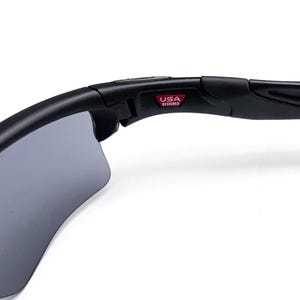 May include: Black sunglasses with gray lenses. The frame is black and features the text "USA DESIGNED" in a red rectangle. The sunglasses have a curved design.