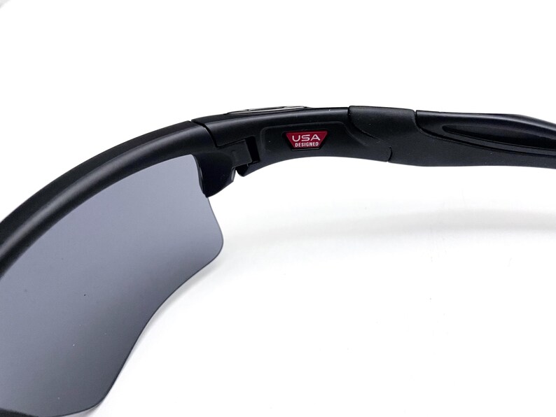 May include: Black sunglasses with gray lenses. The frame is black and features the text "USA DESIGNED" in a red rectangle. The sunglasses have a curved design.