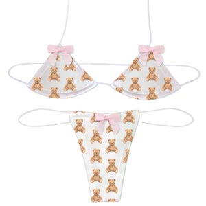 May include: White lingerie set with a repeating pattern of brown teddy bears. The triangle top and thong-style bottom feature pink bows. The set is made of a soft, lightweight material.