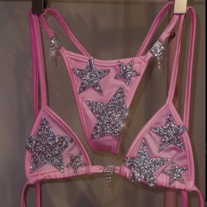 Könnte beinhalten: Pinkfarbener Bikini-Set mit einem Dreiecks-Oberteil und einem hochgeschnittenen Unterteil. Beide Teile sind mit silbernen Glitzersternen verziert.