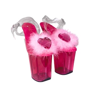 May include: A pair of hot pink platform heels with clear straps. Each heel features a fluffy pink trim, a heart-shaped jewel, and a clear, cylindrical heel. The shoes are set against a white background.