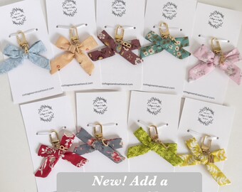 Peut inclure: Divers assortiments de porte-clés en forme de nœud papillon, aux couleurs et motifs variés, dont des motifs floraux et unis. Chaque porte-clés est doté d'un fermoir doré et est fixé à une carte blanche. Le texte "New! Add a Monogram Charm!" est affiché.