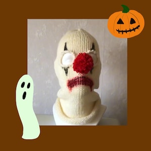 May include: White knitted balaclava with a red pom-pom nose and black eyes, resembling a clown. The balaclava has a red painted mouth and a white background.