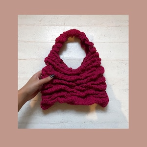 May include: A bright pink crocheted tote bag with a slouchy, relaxed design. The bag has a single handle and a wide, open top.
