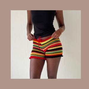 May include: Striped crochet shorts in red, yellow, green, and black with a red drawstring tie. The shorts are worn by a person with a black tank top.