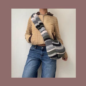 May include: A striped knitted bag worn as a crossbody bag. The bag is made of grey, black, white, and green yarn. The person wearing the bag is wearing a brown corduroy jacket and blue jeans.