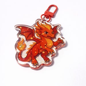 Puede incluir: Un llavero de dragón rojo con un cierre rojo. El dragón tiene alas naranjas y rojas, una cola de fuego y detalles de estrellas. El dragón está sobre un fondo transparente en forma de nube.