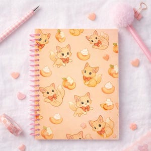 May include: A spiral-bound notebook with a peach-colored cover featuring a repeating pattern of cartoon kittens with bows, flowers, and peaches. The notebook has a pink spiral binding and is surrounded by pink accessories.