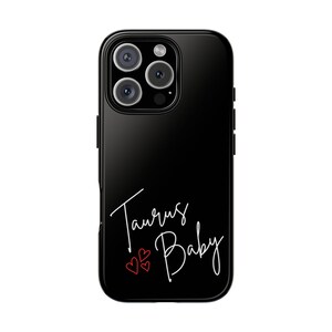 May include: Black phone case with a glossy finish. The case features the words "Taurus Baby" in white script, with three small red hearts below. The phone case has a camera module with three lenses.