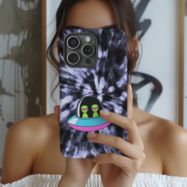 K&ouml;nnte beinhalten: Smartphone-H&uuml;lle mit einem Batikmuster in Schwarz und Lila. Die H&uuml;lle zeigt eine Cartoon-Illustration eines UFOs mit zwei gr&uuml;nen Au&szlig;erirdischen. Das UFO hat einen blauen und rosa Rand.