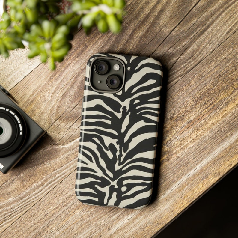 K&ouml;nnte beinhalten: Eine Handyh&uuml;lle mit Zebra-Muster in Schwarz und Wei&szlig; f&uuml;r ein iPhone. Die H&uuml;lle hat eine erh&ouml;hte Kamera-Erhebung mit zwei Objektiven.