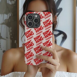 May include: White iPhone case with a repeating pattern of red "DUM DUMS CHERRY" text. The case has a black camera lens.