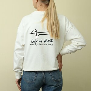 May include: White long-sleeve sweatshirt with a black line drawing of a dachshund on the back. The text "Life is short but my doxie is long" is printed below the dachshund.