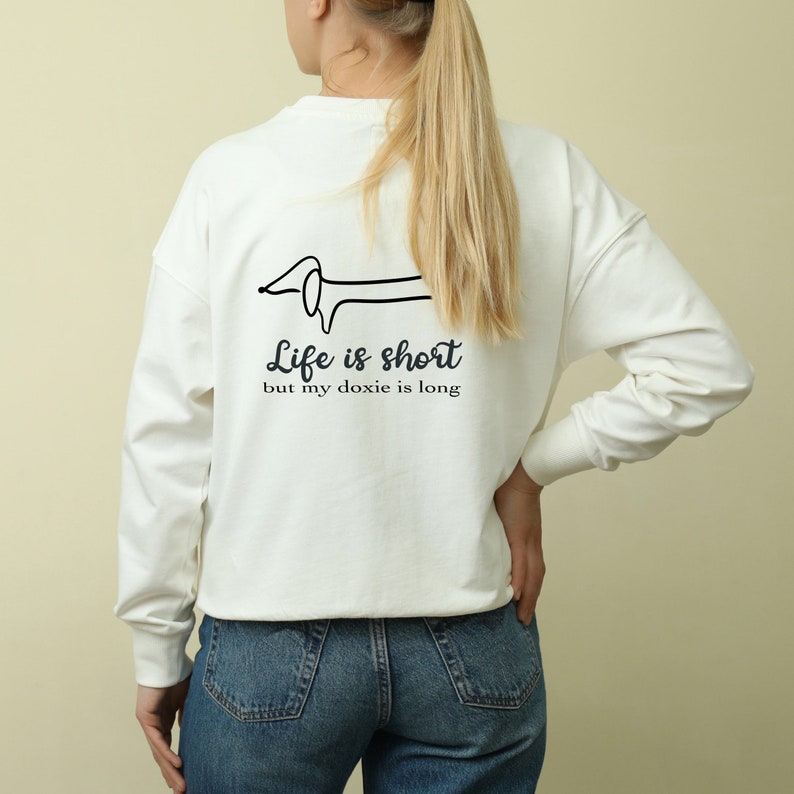 May include: White long-sleeve sweatshirt with a black line drawing of a dachshund on the back. The text "Life is short but my doxie is long" is printed below the dachshund.