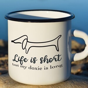 May include: White enamel mug with a black rim and handle. It features a black line drawing of a dachshund and the text "Life is short but my doxie is long."