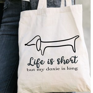 May include: White canvas tote bag with a black line drawing of a dachshund and the text "Life is short but my doxie is long".