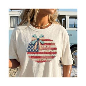Puede incluir: Camiseta color crema con un diseño patriótico. El diseño presenta una bandera estadounidense desgastada con un lazo azul. La bandera incorpora estrellas, rayas y patrones florales en rojo, blanco y azul.