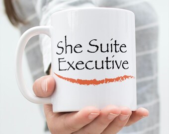 Pode incluir: Caneca de cerâmica branca com um design preto e laranja. A caneca diz "She Suite Executive" com uma linha laranja-avermelhada abaixo do texto.