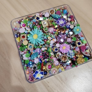 May include: A clear plastic container filled with colorful embellishments, including flowers, rhinestones, and beads. The container is on a wooden surface.