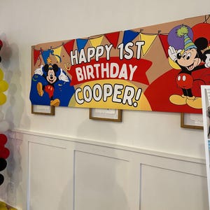 Puede incluir: Una pancarta de cumplea&ntilde;os con el texto "HAPPY 1ST BIRTHDAY COOPER!" y dibujos animados de Mickey Mouse. La pancarta tiene un fondo marr&oacute;n con detalles en rojo, amarillo y azul. Cuelga de una pared blanca.