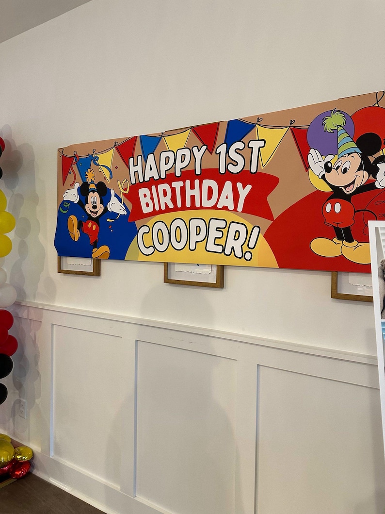 Puede incluir: Una pancarta de cumplea&ntilde;os con el texto "HAPPY 1ST BIRTHDAY COOPER!" y dibujos animados de Mickey Mouse. La pancarta tiene un fondo marr&oacute;n con detalles en rojo, amarillo y azul. Cuelga de una pared blanca.