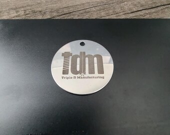 May include: A silver round keychain with a black engraved logo that says "tdm Triple D Manufacturing".