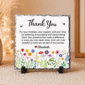 Può includere: Una targa quadrata in ardesia con bordi grezzi e cesellati che riporta il testo Thank You, seguito da un messaggio di gratitudine e il nome Elizabeth con un cuore rosso. Il design include fiori di campo colorati ad acquerello e due farfalle.
