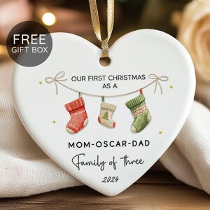 K&ouml;nnte beinhalten: Weihnachtsornament in Herzform mit goldenem Band. Das wei&szlig;e Keramikornament tr&auml;gt den Text "OUR FIRST CHRISTMAS AS A" und "MOM-OSCAR-DAD Family of three 2024". Drei Str&uuml;mpfe sind in Rot, Wei&szlig; und Gr&uuml;n dargestellt.