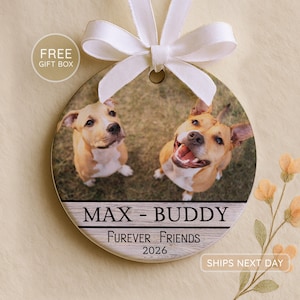 May include: A round ceramic ornament featuring a photograph of two tan and white dogs. Below the photo, the text reads MAX - BUDDY, FUREVER FRIENDS, and 2026 on a wood-grain background. A white ribbon is tied in a bow at the top of the ornament.