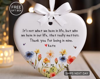 Op de afbeelding: Een wit hartvormig ornament met een bloemenontwerp en een wit lint. Het ornament heeft de tekst "Het is niet wat we in het leven hebben, maar wie we in ons leven hebben, dat er echt toe doet. Bedankt dat je in de mijne bent. Kate."