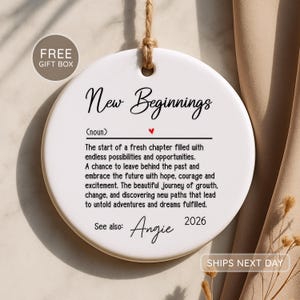 Könnte beinhalten: Ein rundes, weißes Keramikornament, das an einer Juteschnur hängt. Der Text lautet New Beginnings (noun), gefolgt von einer Definition über Neuanfänge, Wachstum und Veränderung. Es enthält ein kleines rotes Herz, den Namen Angie und das Jahr 2026.
