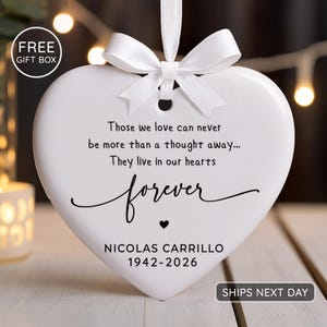 Può includere: Un ornamento in ceramica bianco a forma di cuore con un fiocco di nastro bianco sulla parte superiore. Il testo recita: Those we love can never be more than a thought away... They live in our hearts forever, seguito dal nome NICOLAS CARRILLO e 1942-2026.