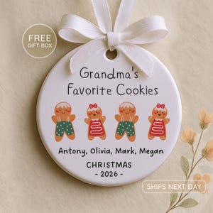 Peut inclure: Une d&eacute;coration ronde en c&eacute;ramique blanche avec un ruban blanc sur le dessus. Le texte indique Grandma's Favorite Cookies, suivi de quatre figurines en pain d'&eacute;pices, des noms Antony, Olivia, Mark, Megan et CHRISTMAS - 2026.